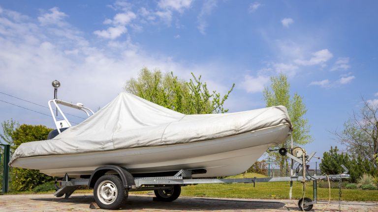 Big modern inflatable motorboat ship covered with grey or white protection tarp standing on steel semi trailer at home backyard on bright sunny day with blue sky on background. Boat vessel storage.