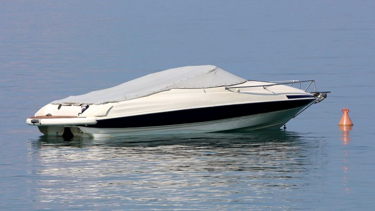 White speed boat covered with light grey tarpaulin protection anchored in local harbor next to orange buoy surrounded with calm clear blue sea on warm sunny summer day